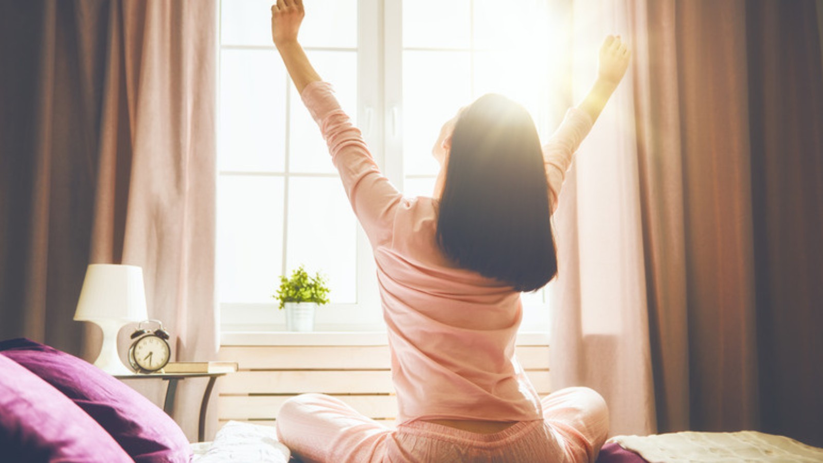 Femme s'étirant au réveil, assise sur son lit devant une fenêtre ensoleillée